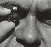 A black and white close up of Aboriginal photographer Mervyn Bishop face. He is holding a tiny camera up to one eye.
