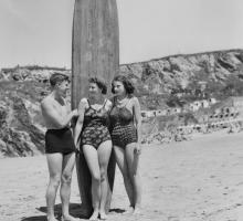 A black and white photo of a man and two women standing in front of a surfboard on the beach.