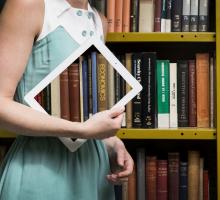 woman walks past a library stack with an ipad showing the books on the stack behind