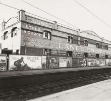 A black and white image of a large two-storey building next to train tracks. The building is covered in advertising signage with the largest sign for Lane's Emulsions, spanning across the whole building. 