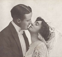 A couple from the 1920's dressed in formal attire standing face to face with their noses touching.